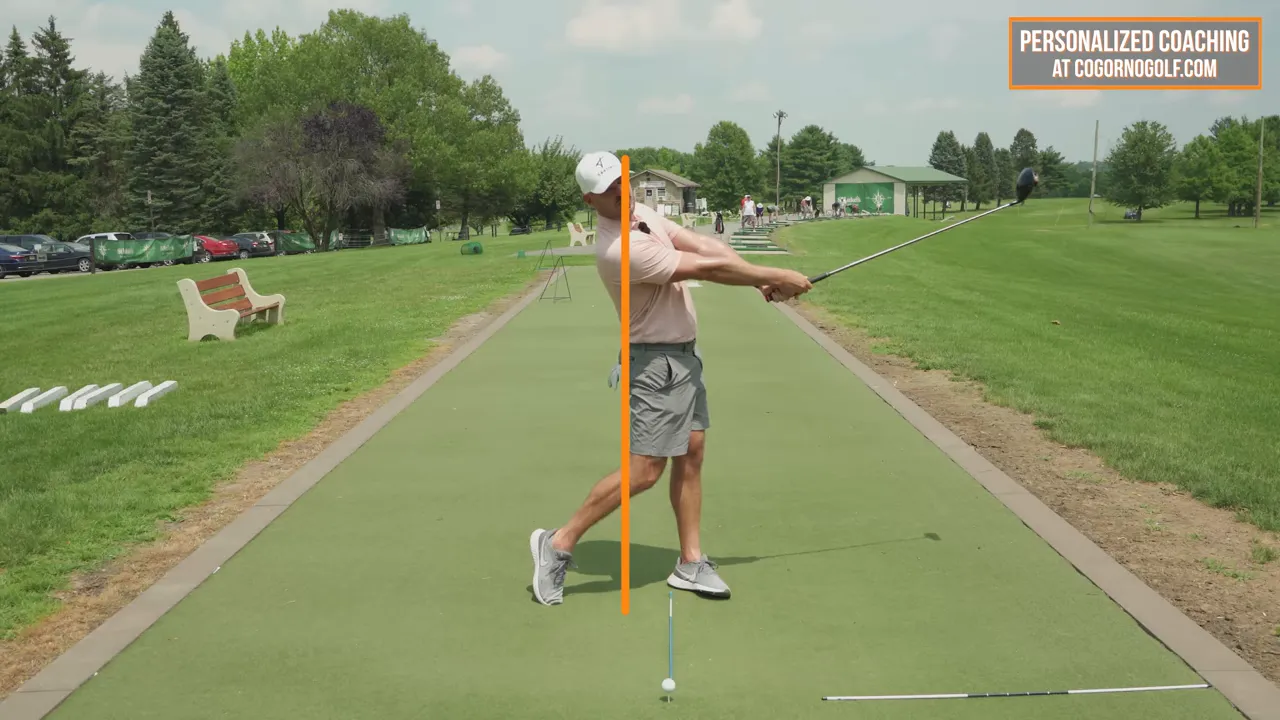 Golfer in full follow-through on a driving range with an orange vertical line showing the head slightly behind the line and the club extended away