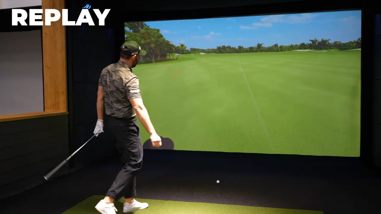 Instructor walking away while the simulator displays the ball flight replay and trajectory across the screen after the shot.
