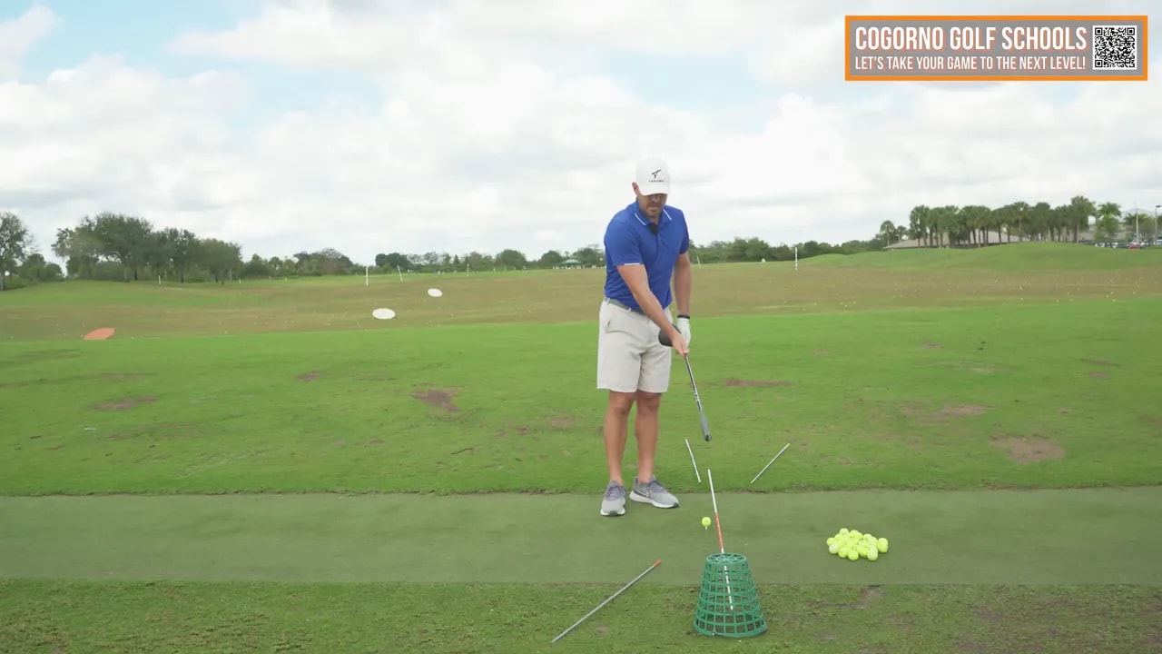 Coach addressing the tee with an alignment stick and training basket visible, demonstrating inside-to-out setup and tilt