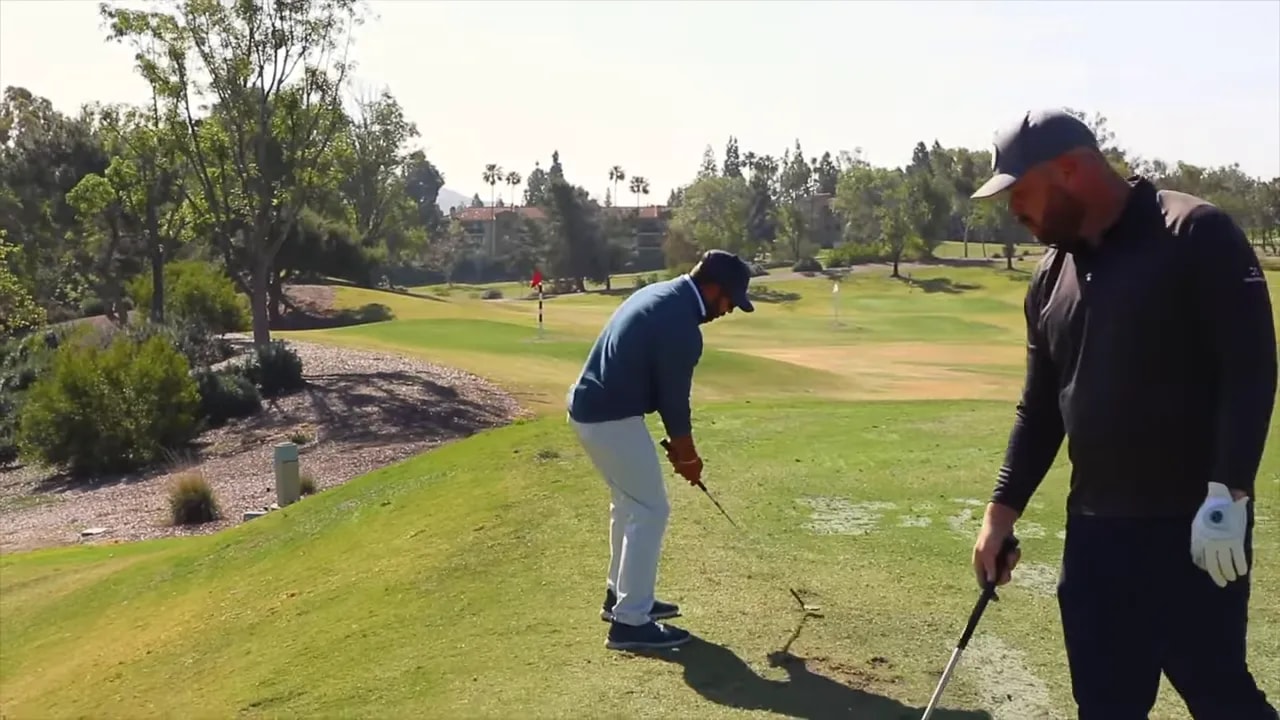 Player striking a wedge with a visible divot in front of the ball while another player observes.