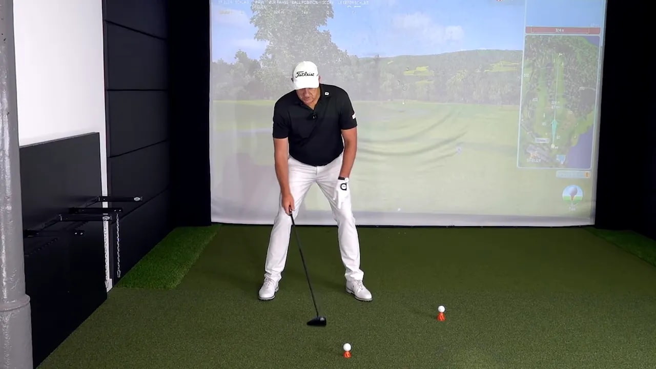 Golf instructor at address in an indoor simulator demonstrating driver setup with ball on tee and balanced stance.
