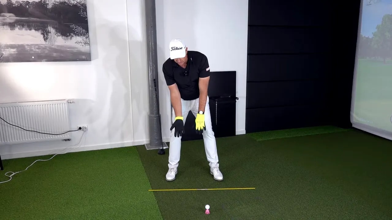 Golf instructor over the ball with hands extended and gloves visible demonstrating the starting hand position for wrist and finger drills.