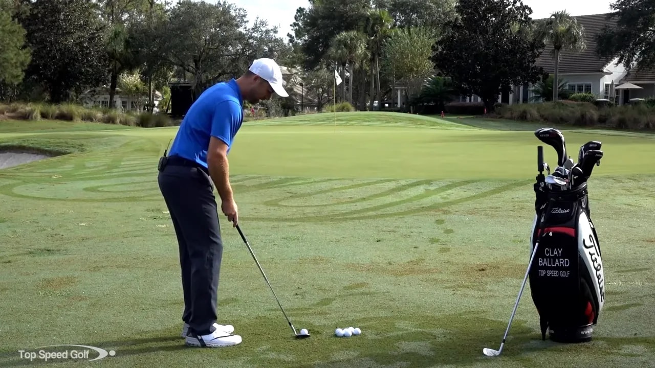 Golfer in pitching setup over golf ball with wedge, golf bag at right and the green and flag in the background.
