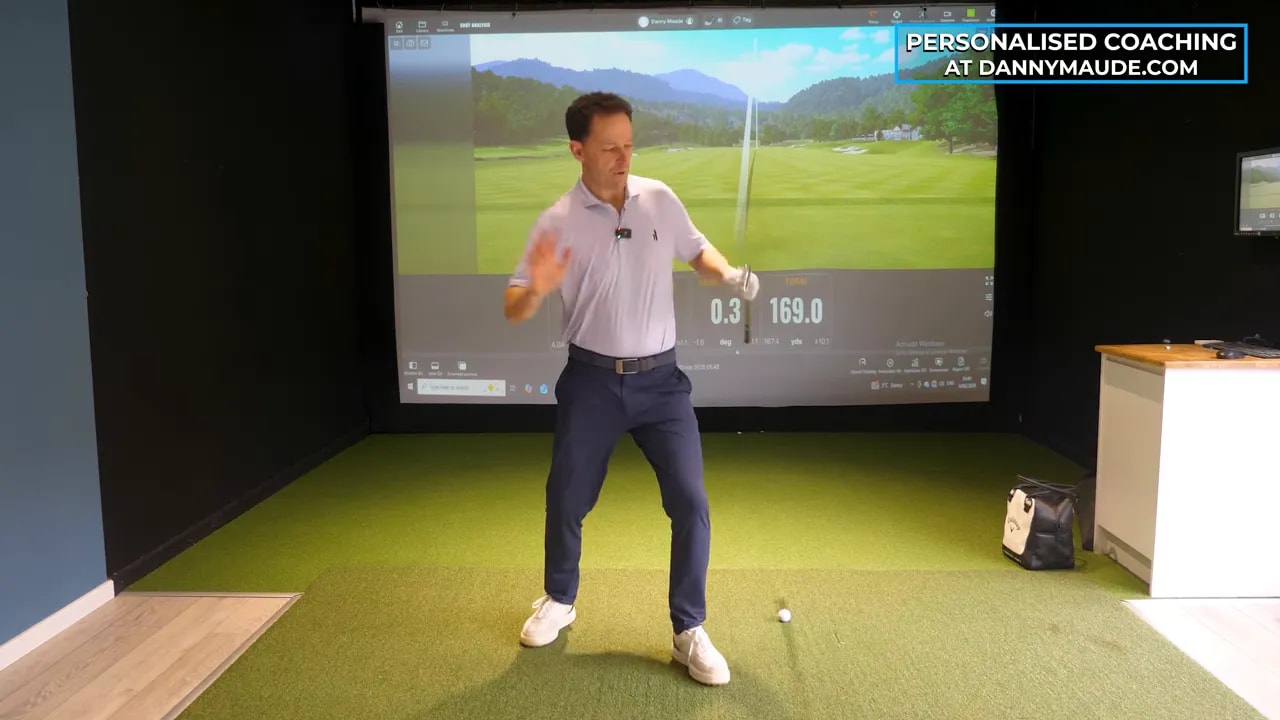 Golf instructor in an indoor simulator demonstrating a lead-foot push and hip rotation while holding a club during a rotational drill