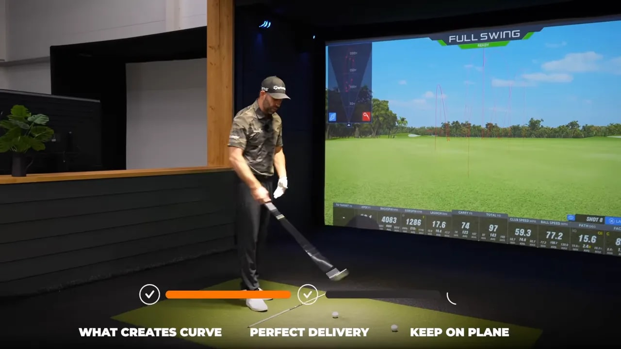 Instructor demonstrating a split‑grip rehearsal beside an alignment stick on the mat with a launch‑monitor screen in the background.