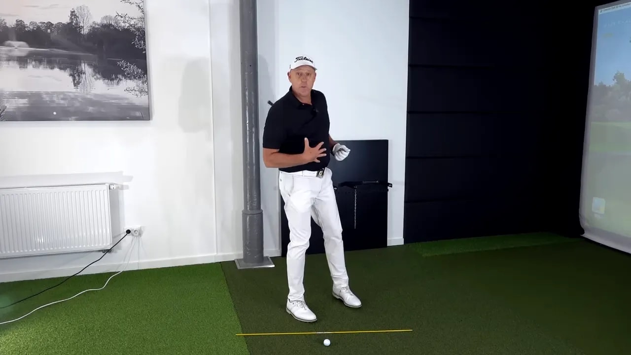 Golf instructor demonstrating the wall setup at address with an alignment stick and ball on the ground in an indoor studio.
