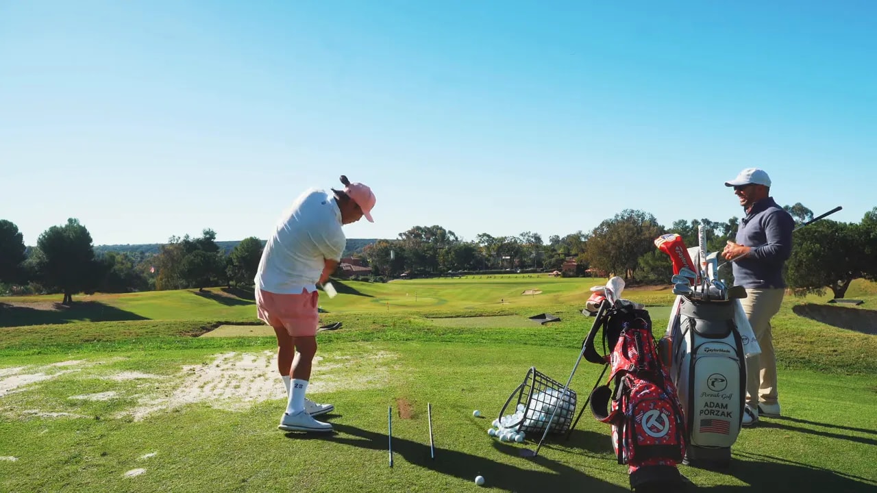 Golfer taking practice shots at a range with a basket of balls and coach nearby
