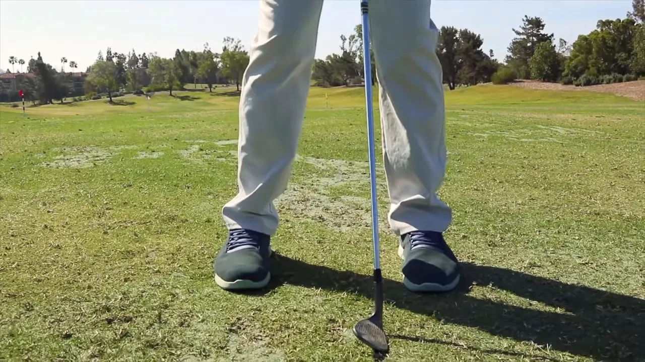 Close-up of a golfer's feet and wedge at address illustrating narrow stance and club position for the door-stop cue.