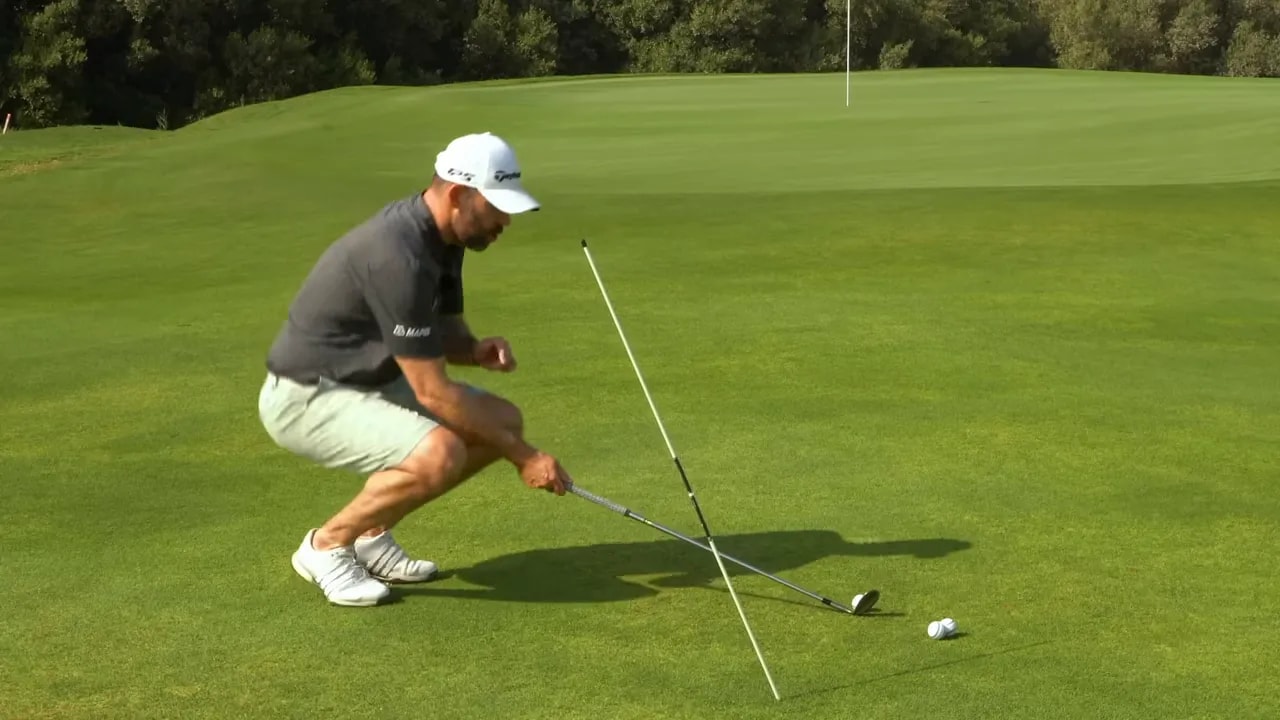 Instructor crouching to place an alignment stick at the clubshaft angle with club and golf ball visible