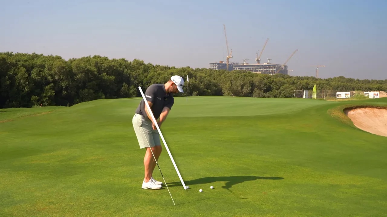 Golfer swinging a pitch with a white line overlay showing the desired vertical swing plane relative to an alignment stick on the ground.