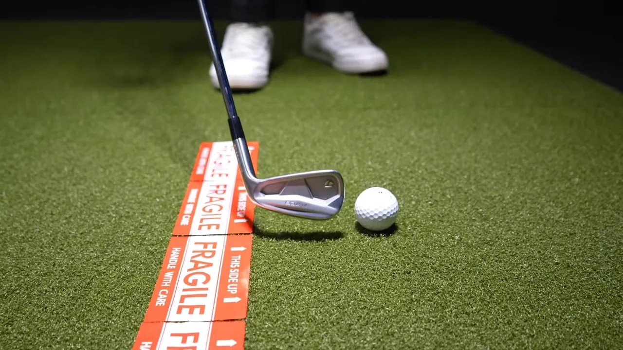 Close-up of an iron next to a golf ball with orange tape on the turf, demonstrating the tape-drill setup.