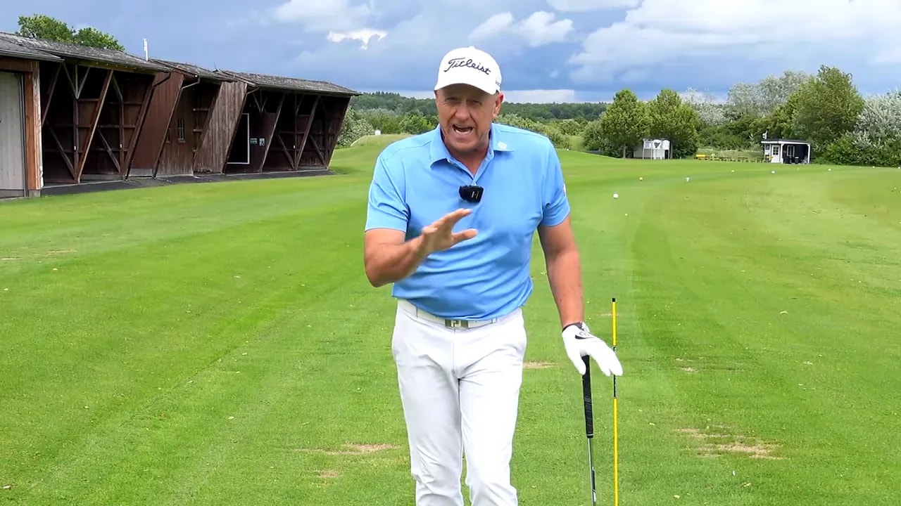 Instructor facing the camera demonstrating troubleshooting tips on the driving range with a club and tour stick visible