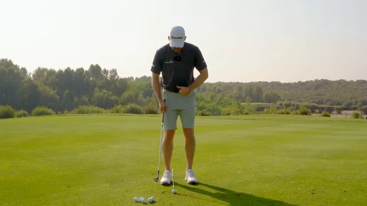 Golf coach demonstrating forward body presence with a hand near his sternum and the ball positioned slightly back in the stance.
