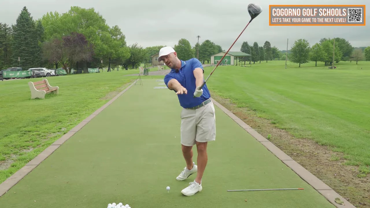 Golf instructor on the driving mat demonstrating a through-impact free release with the club high in the follow-through — full-body view.