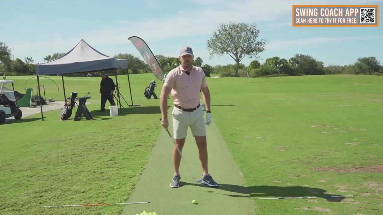 Coach facing the camera at address on a practice mat with alignment rods and a golf ball showing setup position