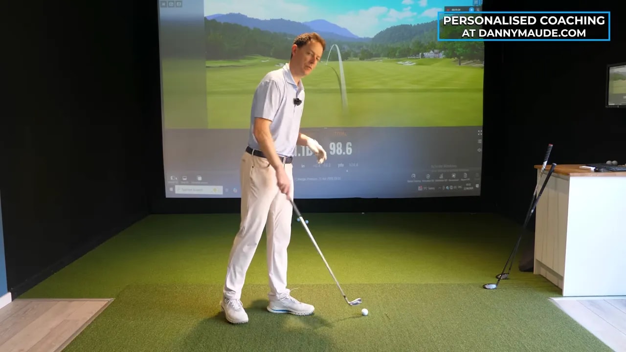 Golf coach pointing toward indoor simulator ball flight and strike feedback metrics on a screen