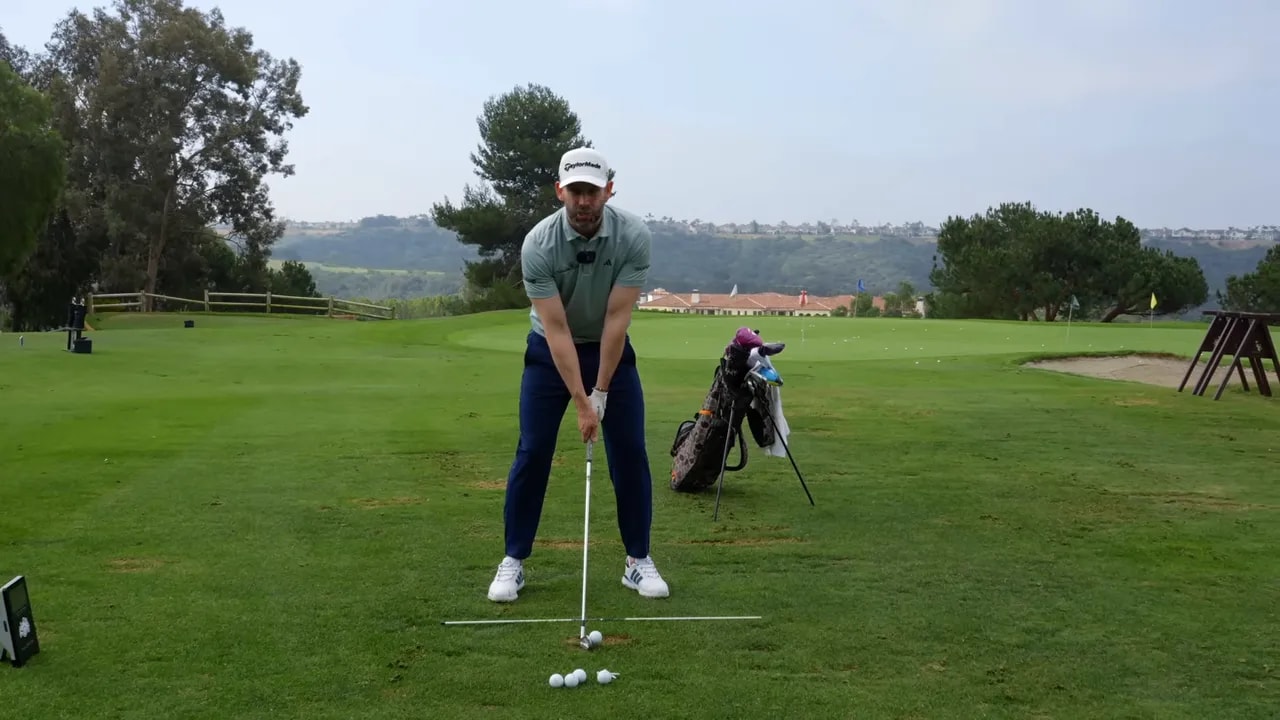 Golf instructor showing full address setup for a compression drill with iron aligned to practice rods on a golf course