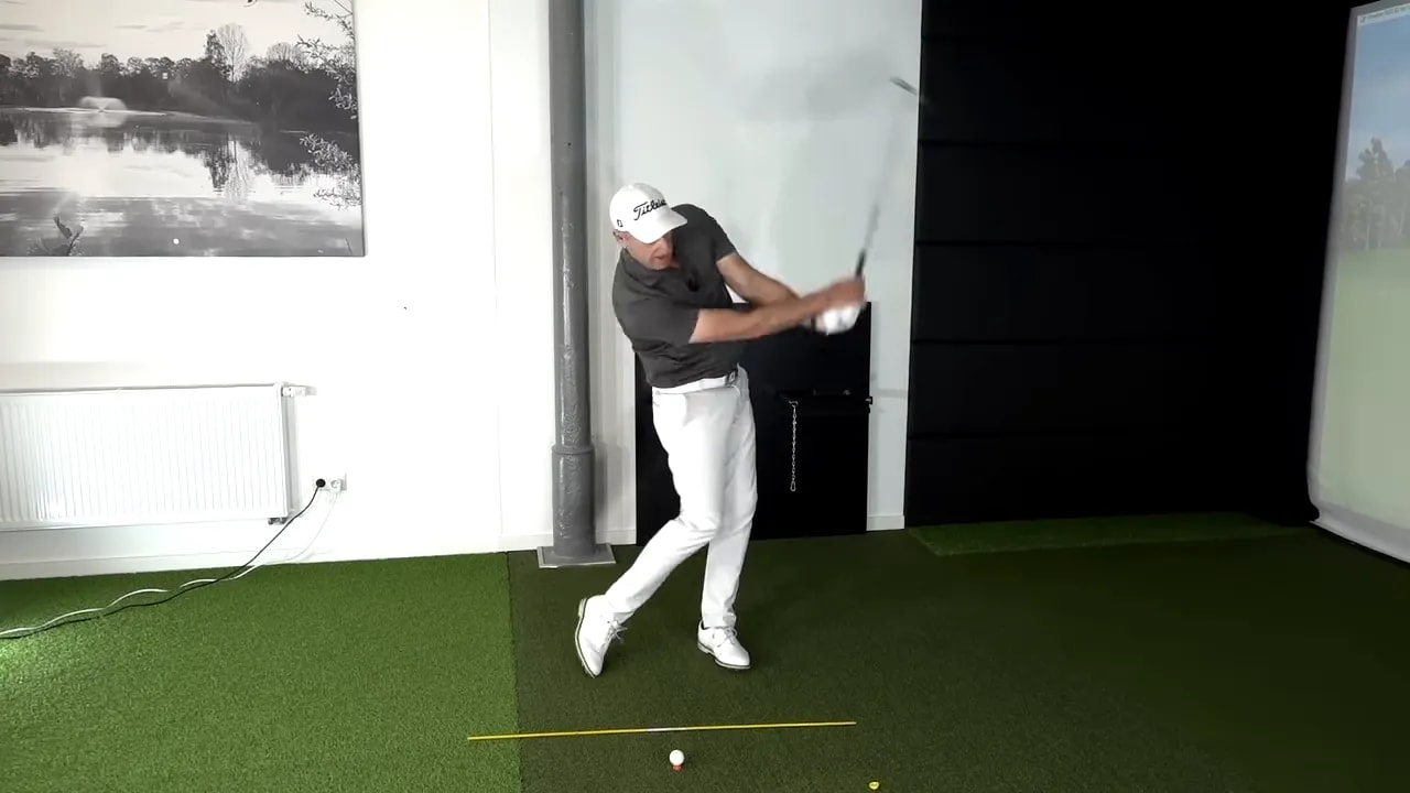 Golfer demonstrating a controlled downswing and strike delivery in an indoor hitting bay with alignment guide line