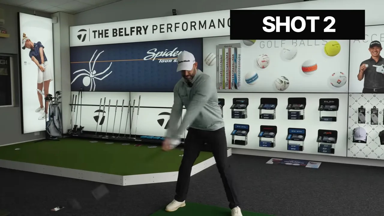 Golfer taking a swing in a fitting bay with the BelFry Performance Lab wall showing the SHOT 2 label