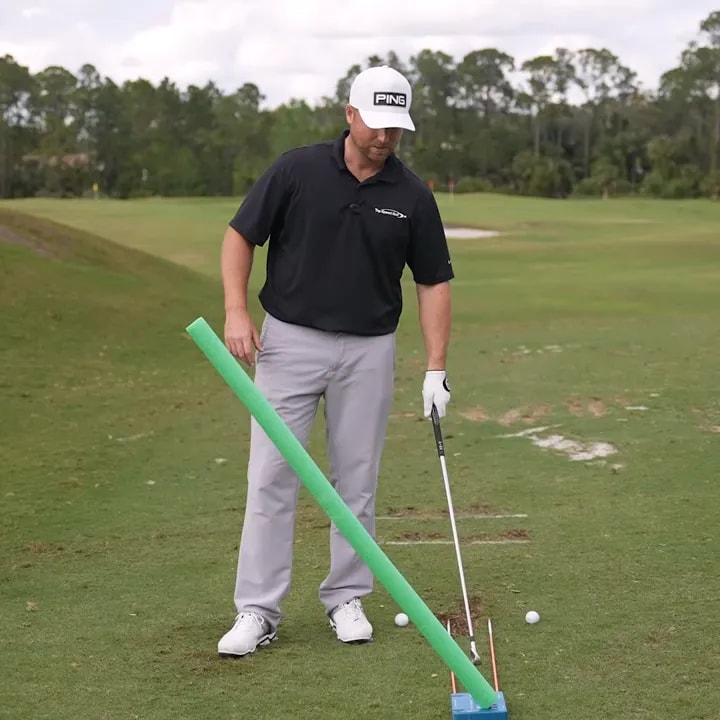 Golfer at address holding a green swing path aid with balls and alignment sticks visible