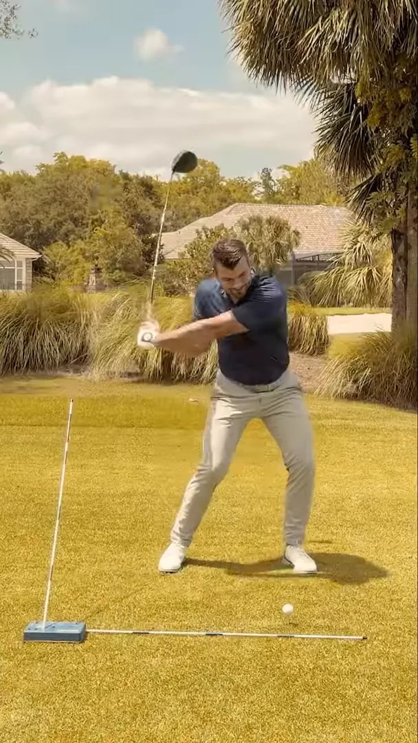 Golfer in downswing with alignment stick on the ground and blue training aid near the ball