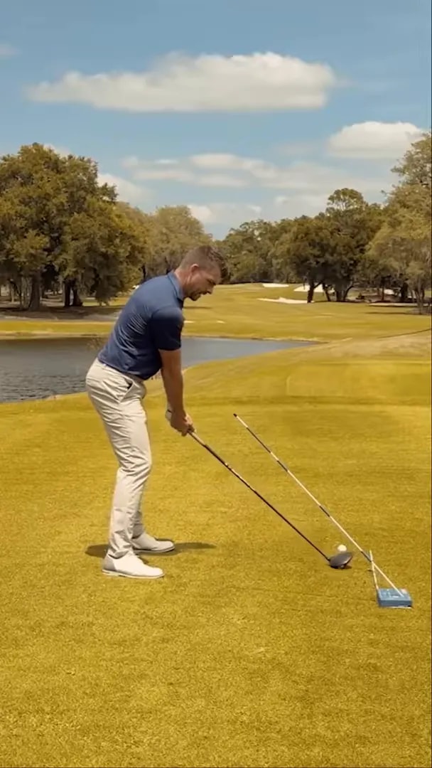 Golfer approaching impact with driver beside an angled alignment stick and blue training aid on the tee box