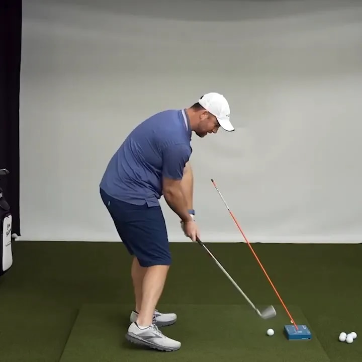 Golfer striking an iron shot while avoiding the angled alignment stick and blue training block
