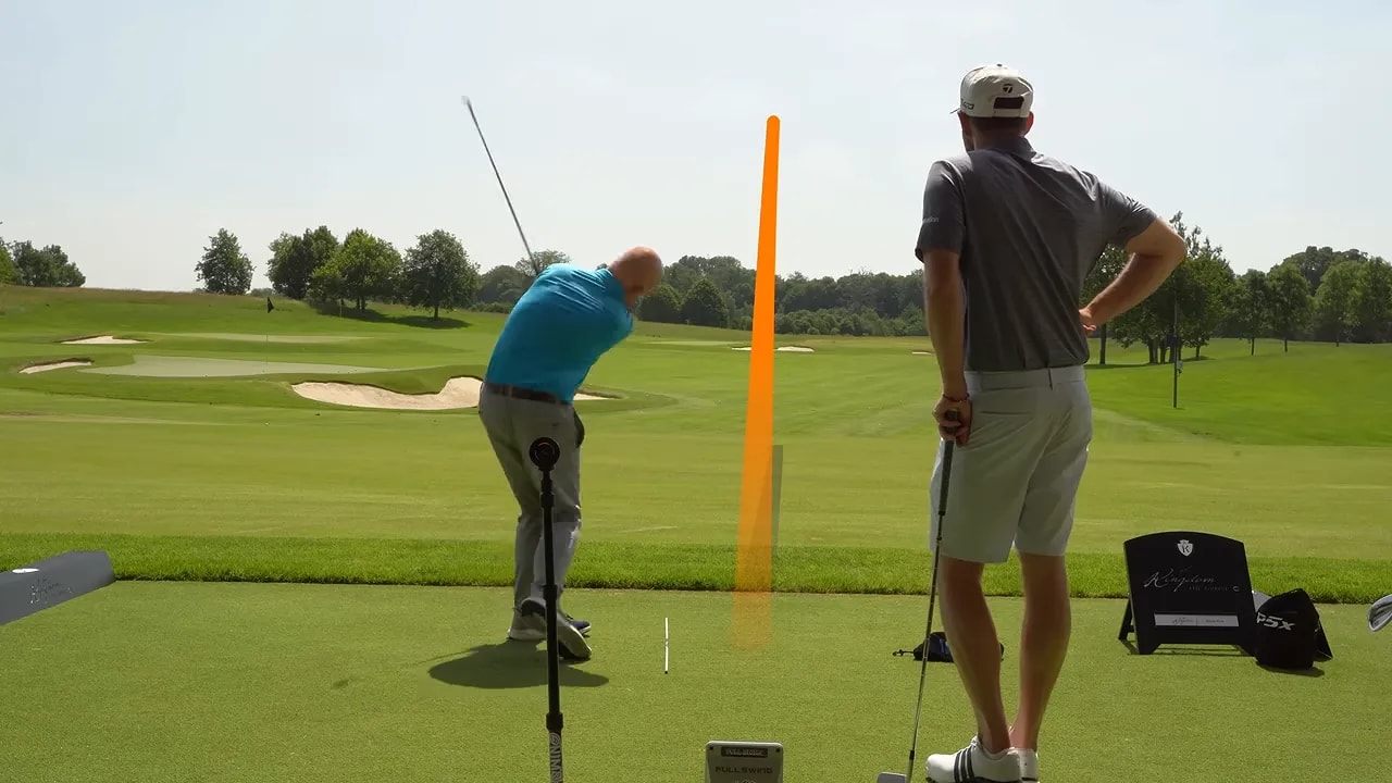 Golfer finishing an iron shot with an orange ball-flight tracer rising ahead