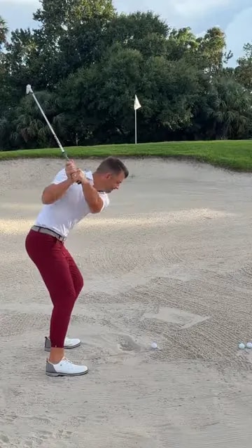 Golfer swinging through a bunker shot with sand spraying toward the green